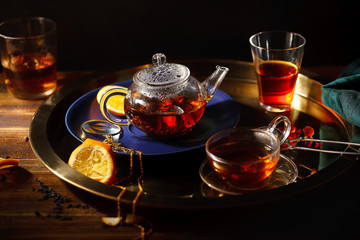 Small glass teapot with hot black tea, cup, glasses, pocket magnifier on golden chain, squeezed orange slice and golden tray on wooden background. Evening light. Overhead view.