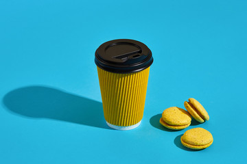 Hot coffee in yellow paper cup with black lid and macaroons on b