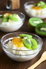 Rice pudding with kiwi pieces, orange jam and cinnamon in small glass bowls, photographed on dark wood with natural light (Selective Focus, Focus on the kiwi piece in the front)
