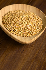 Yellow mustard seeds on small wooden plate, photographed on dark wood with natural light (Selective Focus, Focus on the seeds in the front)