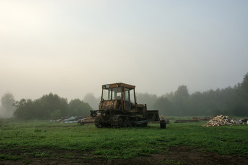 Fototapeta premium The caterpillar tractor stands in the morning fog on the background of logs and trees. Green grass.