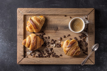 Fototapeta na wymiar Fresh baked traditional croissants and mug of espresso coffee, coffee beans, sugar on wooden tray over black texture background. Top view, space
