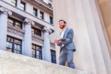 American Businessman with beard, mustache working outside in New York, wearing cadet blue suit,  white undershirt, standing against column on street with vintage buildings, working on laptop computer