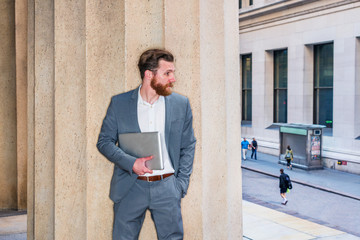 American Businessman with beard, mustache working in New York, wearing cadet blue suit, white undershirt, carrying laptop computer, standing against column outside office building, looking at street..