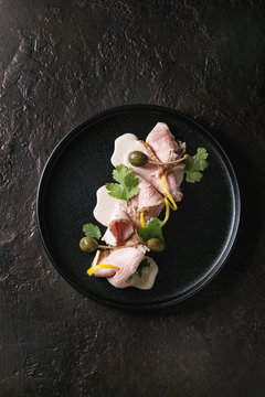 Vitello tonnato italian dish. Thin sliced veal with tuna sauce, capers and coriander served on black plate over dark texture background. Top view, copy space