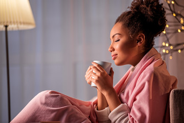 Peaceful african girl is smelling nice coffee aroma from cup. She is sitting on sofa covered by pink blanket. Her eyes are closed with pleasure. Copy space