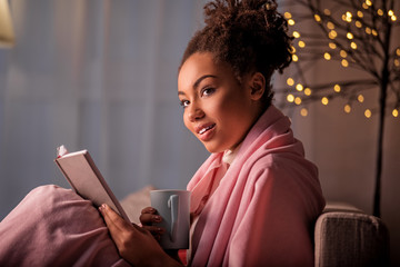 Portrait of cheerful african girl drinking hot beverage at home. She is holding cup and book. Home entertainment concept