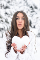 A young woman in a winter forest holds a heart from the snow and sends her kiss.