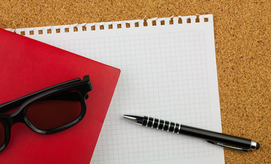 Empty square sheet of white paper wrested from notepad for your text. There are glasses in black thick rim, red notebook, black pen and cork board background.