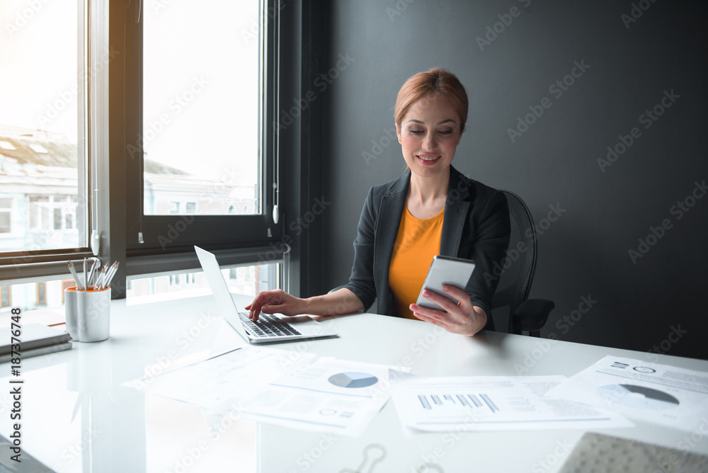 Poster portrait of cheerful businesswoman noting in notebook computer while sending message in mobile. tech