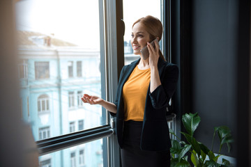 Portrait of happy businesswoman telling by mobile while standing near window in modern apartment. Conversation and labor concept