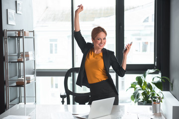 Portrait of cheerful female flourishing arms while holding phone. She locating near table with notebook computer in office. Profession and success entertainment concept