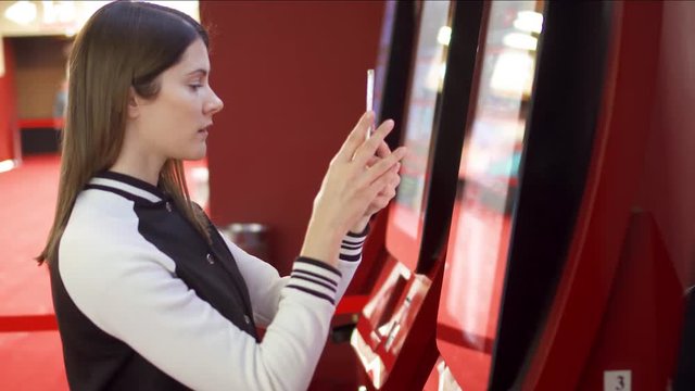 Teenager buying movie ticket from vending machine at movie theater at mall. Woman using mobile to read information about movies online. Making photos of screen, donwloading information from internet