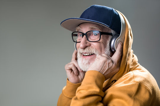 Happy old man with grin from ear to ear. He is wearing headphones over the cap. Copy space in left side. Isolated on grey background