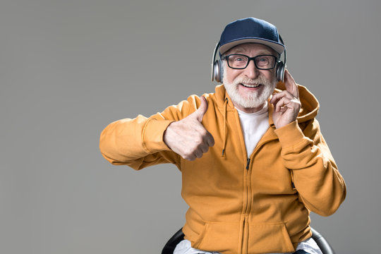 Waist up portrait of satisfied senior sitting on chair and enjoying music. He is looking at camera with broad smile. Isolated on grey background