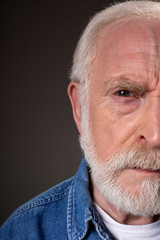 Half face of aging male looking at camera with suspicion. Isolated on grey background. Close up