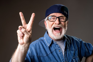 Portrait of cheery greybeard in cap showing peace sign and laughing. Isolated on grey background