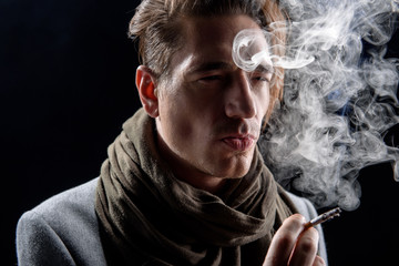 Heavy thoughts. Portrait of pensive young cute man is standing and enjoying his bad habit. He is looking aside dreamily while being in cloud of white smoke. Isolated on black background