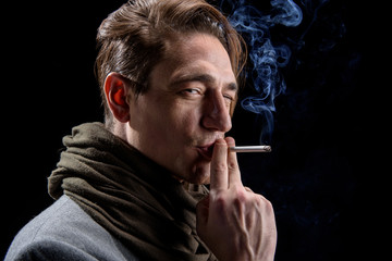 Need relax. Side view of young calm modish man is standing and smoking with pleasure. He is squinting on camera thoughtfully. Isolated on black background