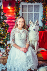 Cute girl in white dress hugging a Husky dog on bench, on the background red house