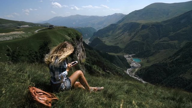 A young blonde girl sits on the grass on a background of mountains with a backpack and takes pictures of the mountains on the phone