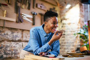 Portrait of a smiling female carpenter sending a voice message with smartphone in her workshop.