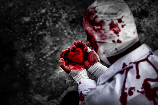 seriously injured man in body wraps bandages bleeding in frustrated depression from unrequited love. holding the red heart smeared with blood in hand. love concept for valentines day. in dark tone.