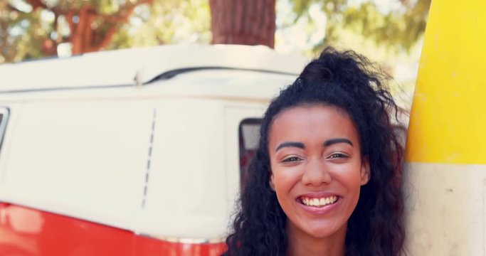 Smiling woman with surfboard standing near camper van 