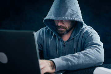 criminal bearded man hides his face under the hood, hacks the password on the laptop