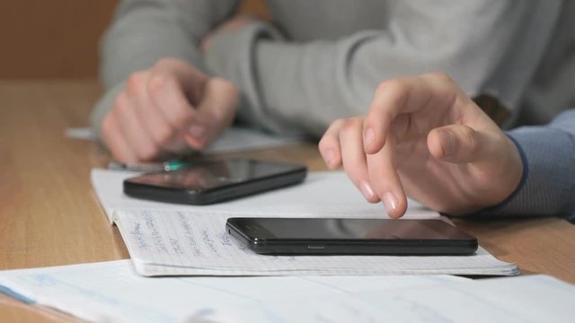 Close-up of hands of two unknown students dressed in shirt sits at desk at lesson and using black smartphones pressing them fingers on the screen and look at the important information.