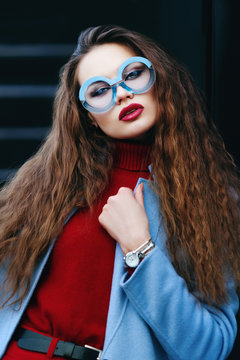 Outdoor portrait of young beautiful fashionable woman posing in street. Model wearing red cashmere turtleneck sweater, blue coat, round sunglasses, wrist watch. Female fashion concept