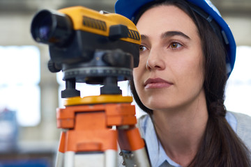 Portrait of female geodesist wearing hardhat looking into optical level mounted on tripod at construction site - obrazy, fototapety, plakaty