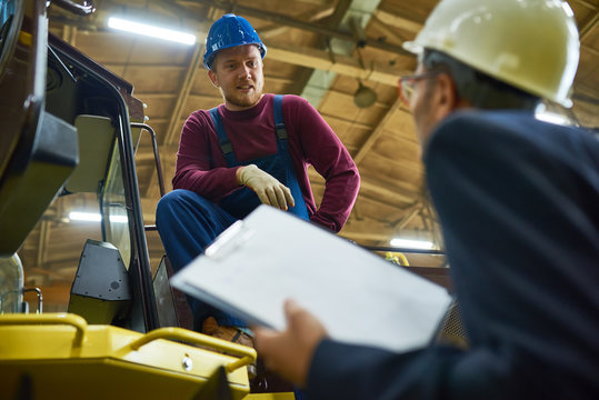 Low angle view of inspector with clipboard in hands asking bearded young worker something while walking along spacious heavy equipment factory