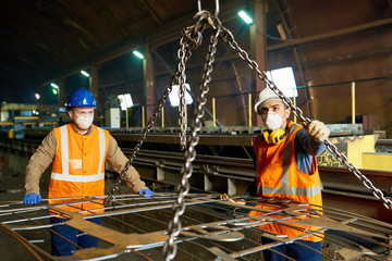 Two young workers of steel plant wearing uniform and filter masks using lifting beam in order to...
