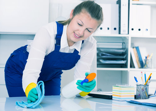 Cheerful female cleaner is cleaning dust from the desk