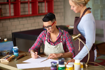 Fototapeta premium Portrait of two modern young women enjoying working together in art studio, sketching draft for new project