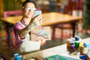 Portrait of modern creative woman taking selfie with her artwork  in art studio, focus oh tattoed hand holding smartphone, copy space
