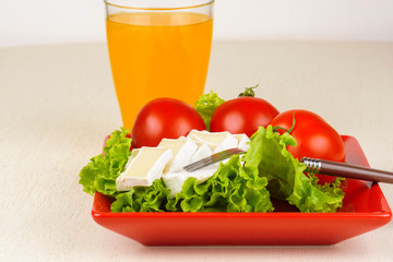 Tomatoes and cheese slices lie on a sheet of fresh salad in a red plate. A glass of orange juice stands side by side on the table.