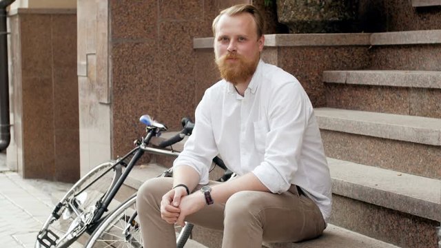 4k footage of stylish red bearded man with vintage bicycle sitting on stone staircase