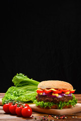 Delicious tasty burger with beef, tomatoes, salad and sauce on wooden table and black background. Space for text or design. 