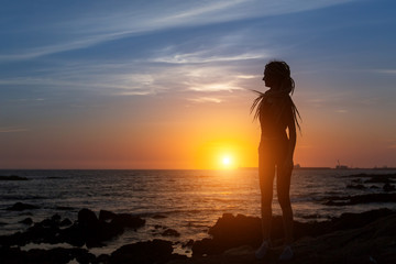 Silhouette of flexible girl with dreadlocks on ocean coast during amazing sunset.