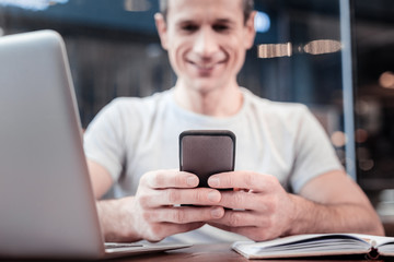 Answer message. Handsome male person leaning arms on the table and keeping smile on his face while typing message