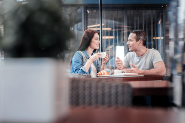 Warm atmosphere. Positive delighted woman sitting opposite her friend and holding cup of coffee in both hands while being in all ears