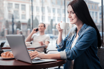Work with pleasure. Amazing young woman sitting in semi position and holding cup near mouth while looking at screen of computer