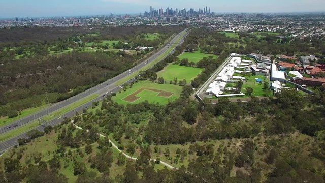Static aerial establishing shot of cars driving on Eastern Freeway, Melbourne Polytechnic and Melbourne CBD skyline in the distance on bright summer day