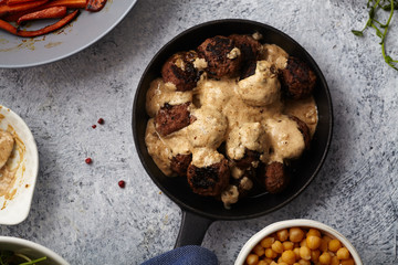 Flatlay with traditional swedish beef meatballs with gravy served in one portion black cast iron pan, handle wrapped in blue towel, plates with sauce, baked carrots and chickpeas. Dinner concept.