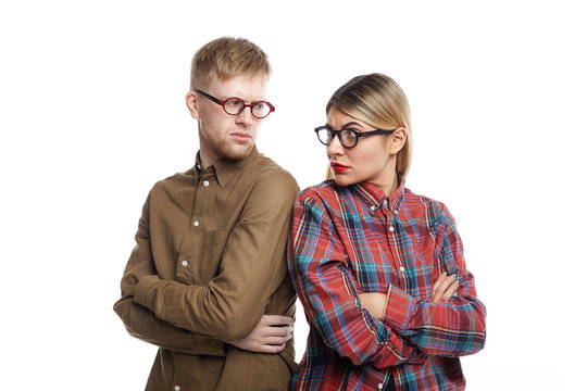 Angry reluctant young blonde woman and unshaven man both in spectacles standing back to back with arms crossed and looking at each other over their shoulders with serious dissatisfied expressions