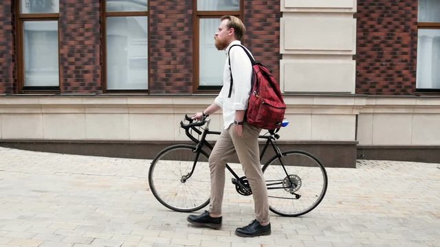 Slow motion video of young bearded man with red bag walking on street with bicyclec