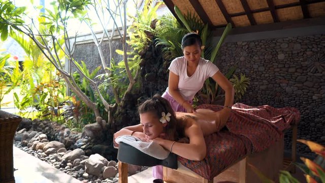 Young caucasian woman with frangipani in ear having outdoor spa massage by balinese masseuse in beautiful garden