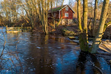 Zelfklevend Fotobehang Spring flood on a small forest river near a red house in the morning sunshine. Fast moving water cascading over rocks and water far beyond the normal riverbanks. Brakne river in Blekinge, Sweden. © imfotograf